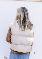 Back view of Model wearing an oatmeal puffer zip-up vest with a slightly cropped fit and pockets.