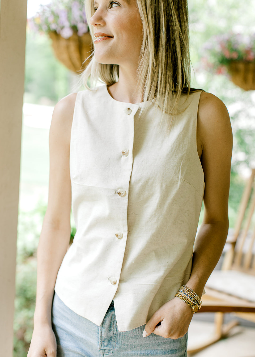 Model wearing a cream sleeveless vest-style top with button front and adjustable tie back.