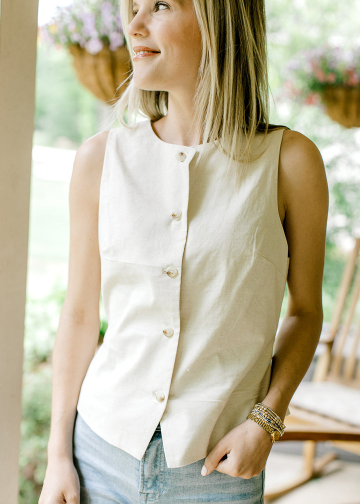 Model wearing a cream sleeveless vest-style top with button front and adjustable tie back.