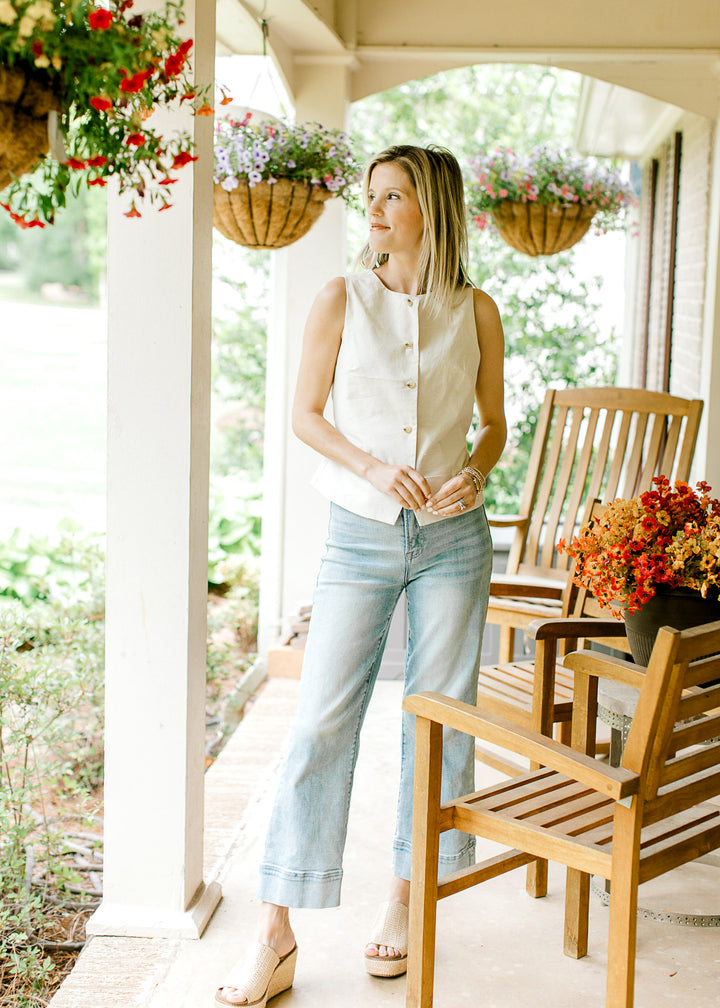 Model wearing jeans and a cream sleeveless vest-style top with button front and tie back.