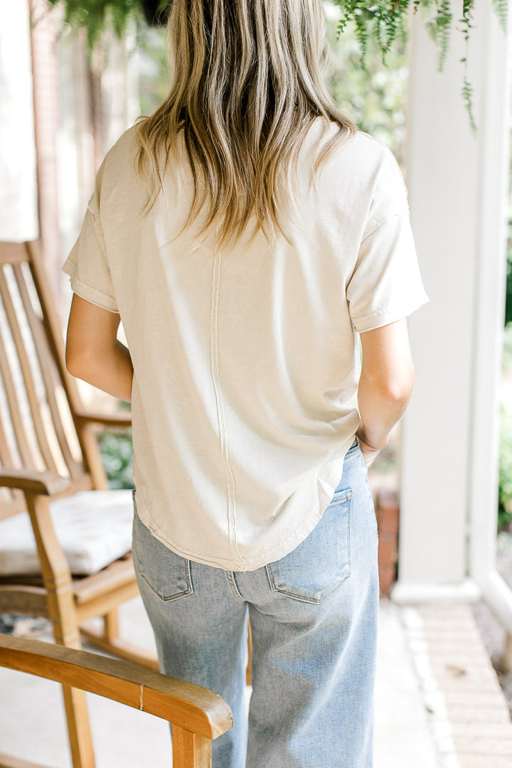 Back view of Model wearing a v-neck oatmeal top with raw edges, short sleeves and a loose fit. 