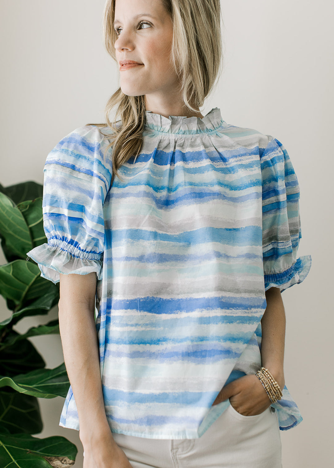 Model wearing a flowy top of varied lines of blue, ruffle detail at neck and short sleeves.