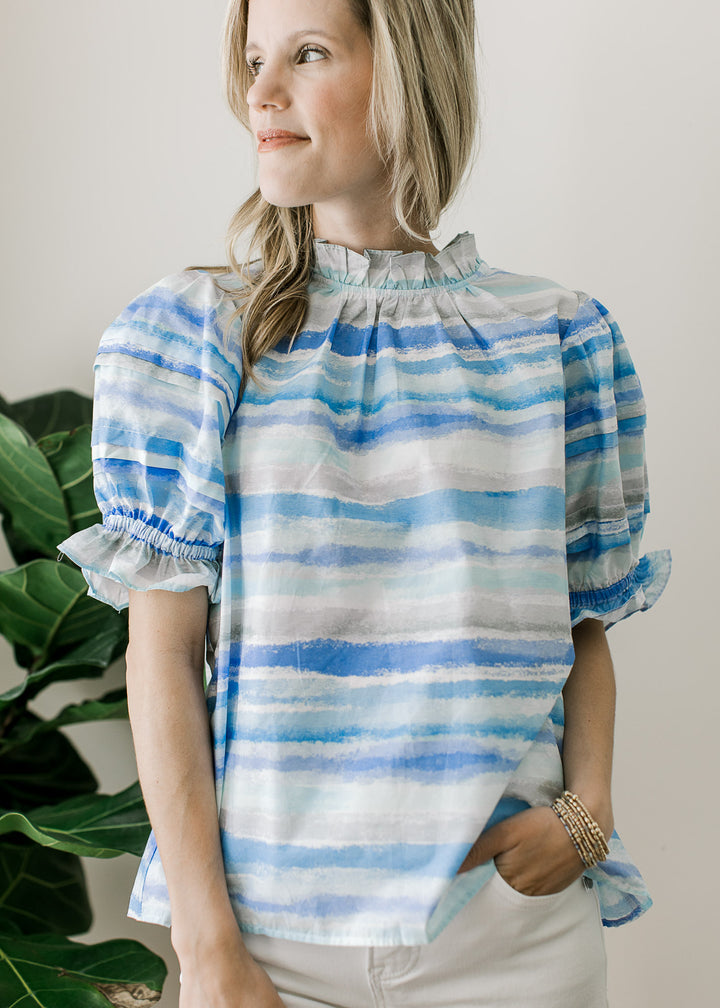 Model wearing a flowy top of varied lines of blue, ruffle detail at neck and short sleeves.