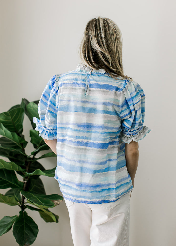 Back view of Model wearing a top of varied shades and lines of blue, keyhole, ruffle detail. 