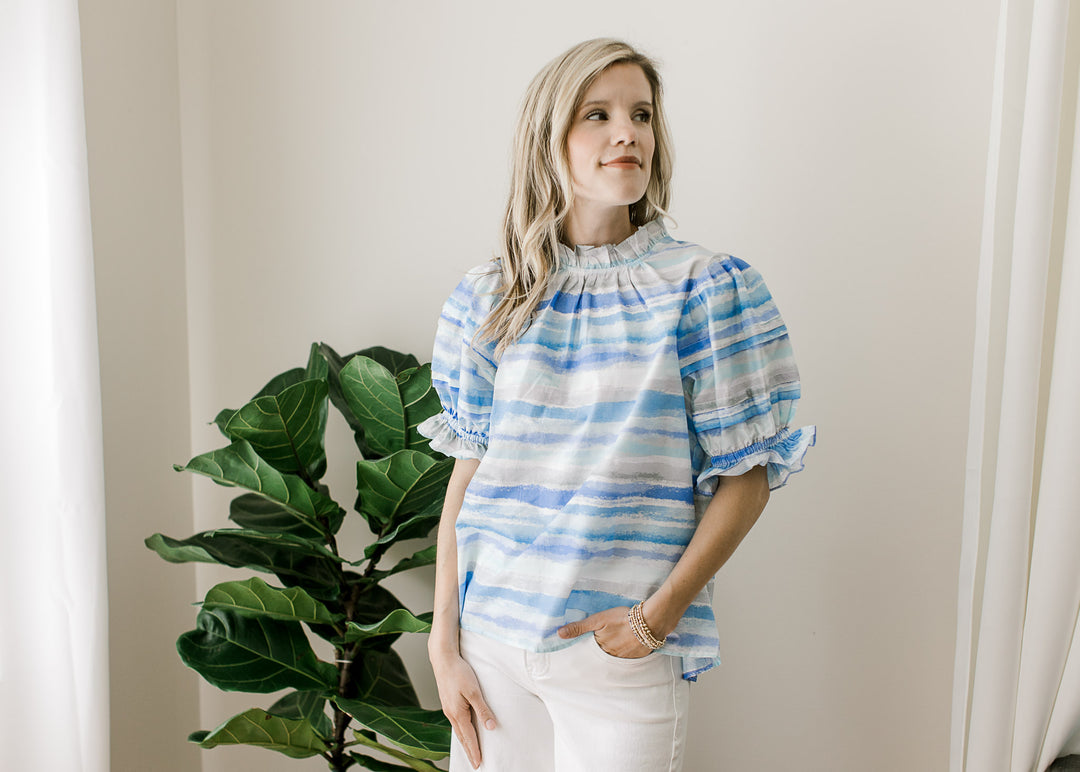 Model wearing white pants and a top of varied shades and lines of blue and bubble short sleeves.