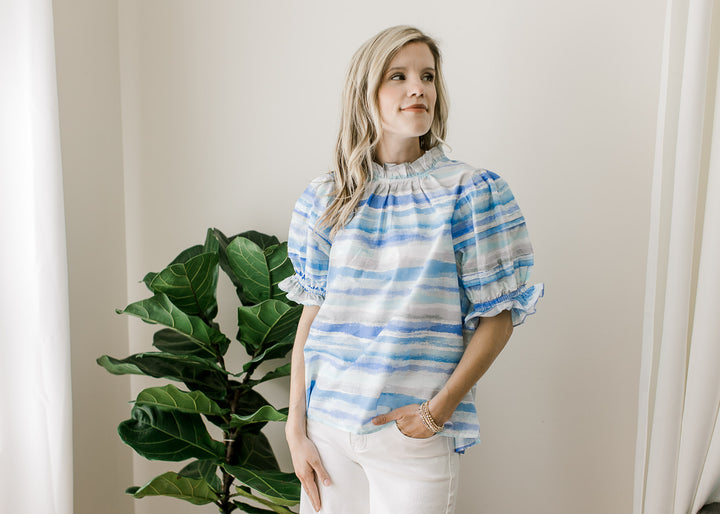 Model wearing white pants and a top of varied shades and lines of blue and bubble short sleeves.