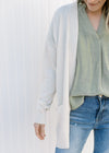 Close up of Model wearing an off white cardigan with an open front, long sleeves and patch pockets.