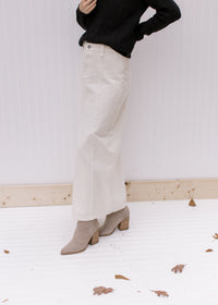Side view of a model wearing a black top and booties with a cream denim skirt with a front slit