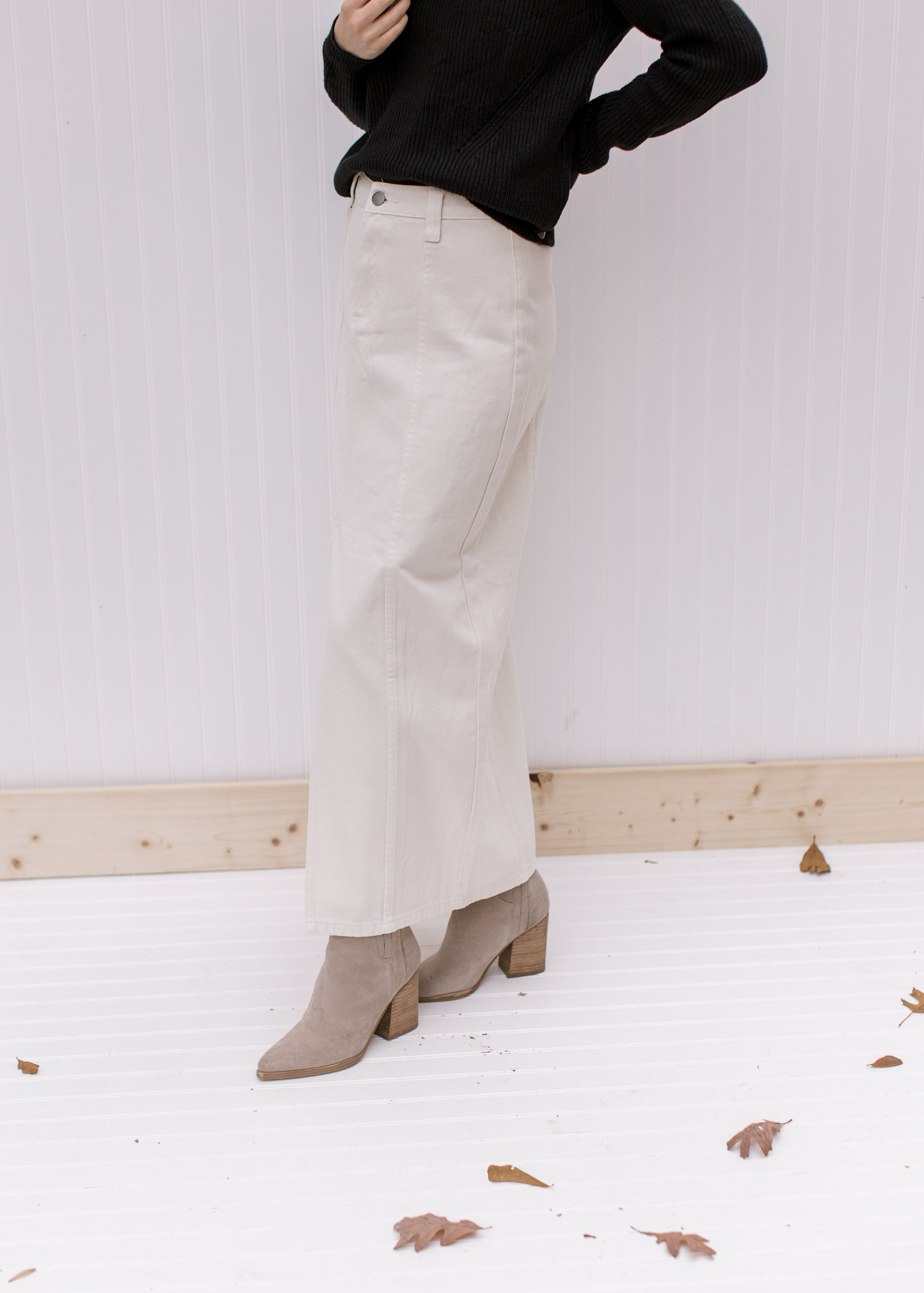Side view of a model wearing a black top and booties with a cream denim skirt with a front slit