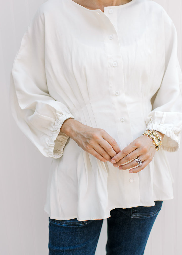 Model wearing an off-white top with button-front, elbow-length sleeves and pleated waistline.