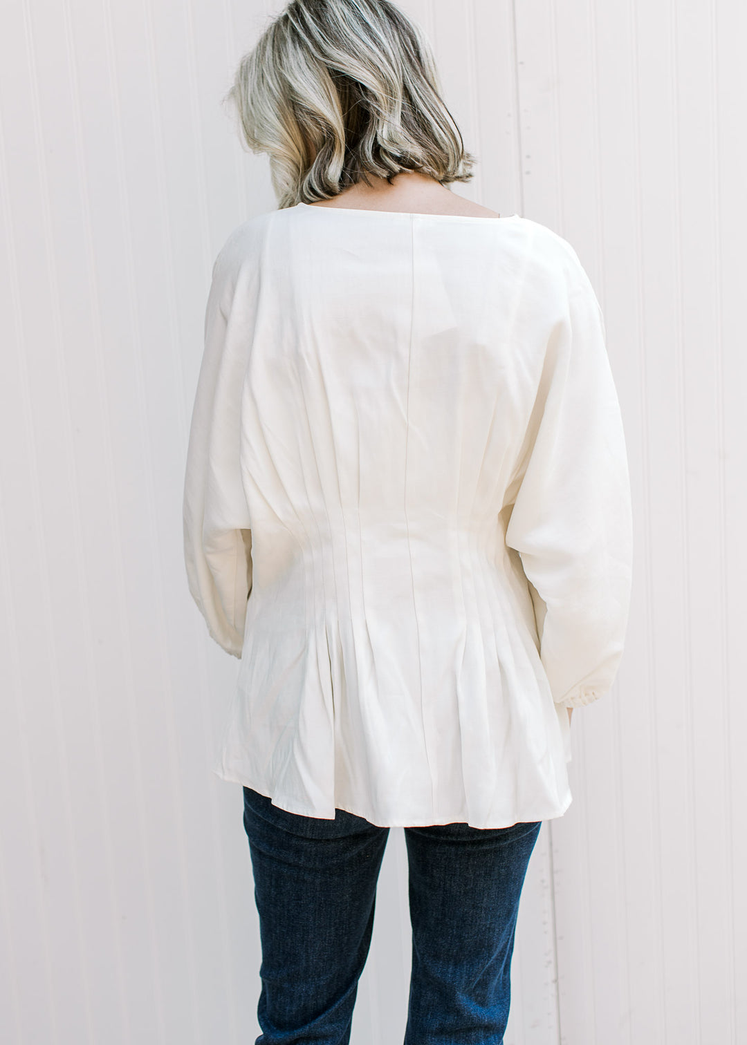 Back view of Model wearing an off-white top with elbow-length sleeves and pleated waistline.