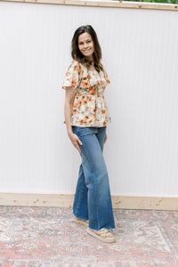 Model wearing jeans, sandals and a cream top with pink floral pattern and short sleeves.