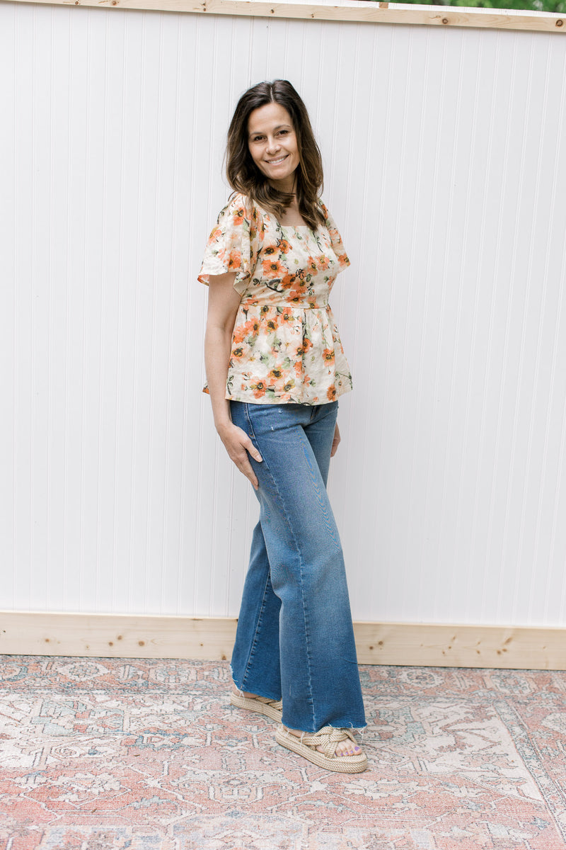 Model wearing jeans, sandals and a cream top with pink floral pattern and short sleeves.