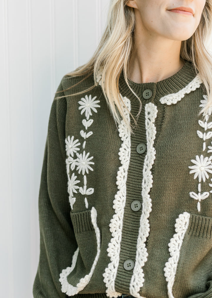 Close up of cream embroiled floral and lace on an olive button up cardigan with pockets. 