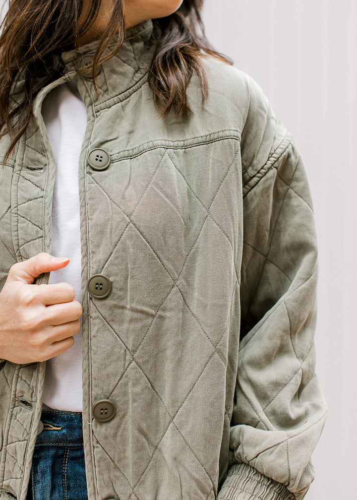 Model wearing an olive quilted jacket with button front, long sleeves, pockets and a mock neckline. 