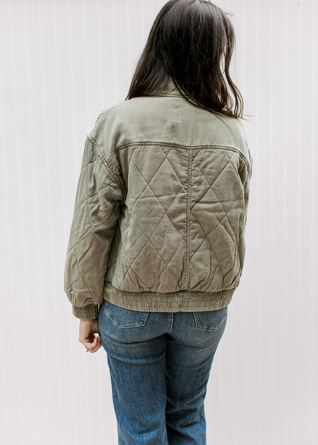 Back view of Model wearing an olive quilted jacket with long sleeves, pockets and a mock neck. 