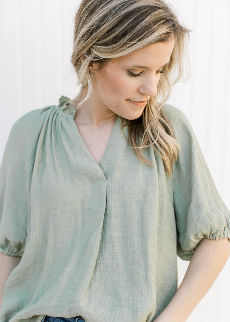 Model wearing a sage flowy top with ruffles at the neck and a pleat down the front. 