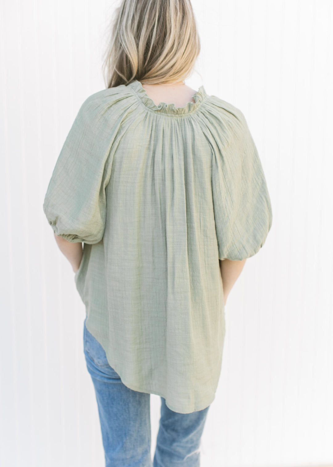 Back view of Model wearing a sage flowy top with ruffles at the neck and a pleat down the front.