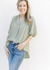 Model wearing jeans with a sage flowy top with ruffles at the neck and a pleat down the front.