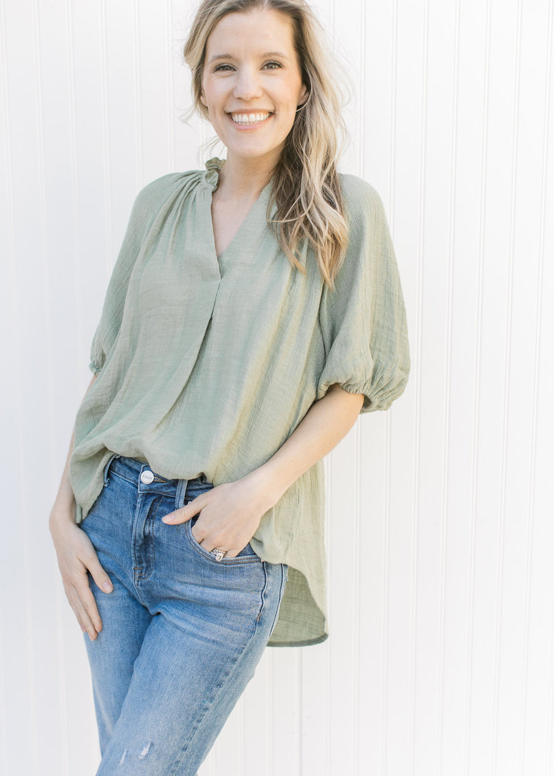 Model wearing jeans with a sage flowy top with ruffles at the neck and a pleat down the front.