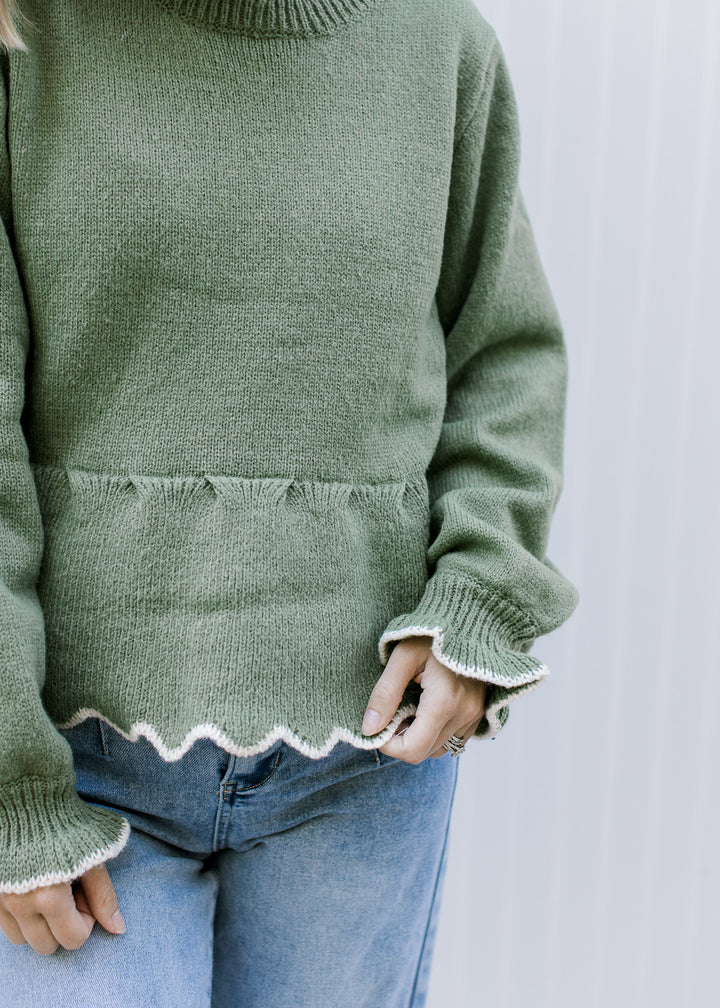 Close up of detail at peplum fit and cream scalloping at hem and cuff of an olive sweater.  