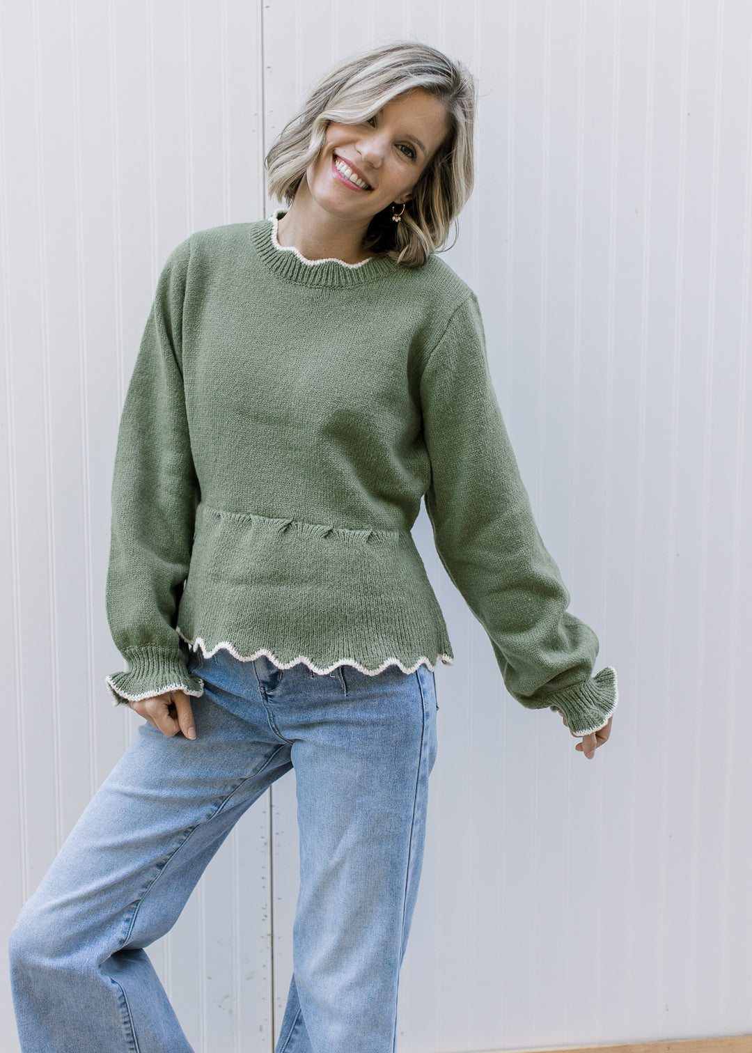 Model wearing an olive peplum sweater with cream scalloped trim, round neck and long sleeves.