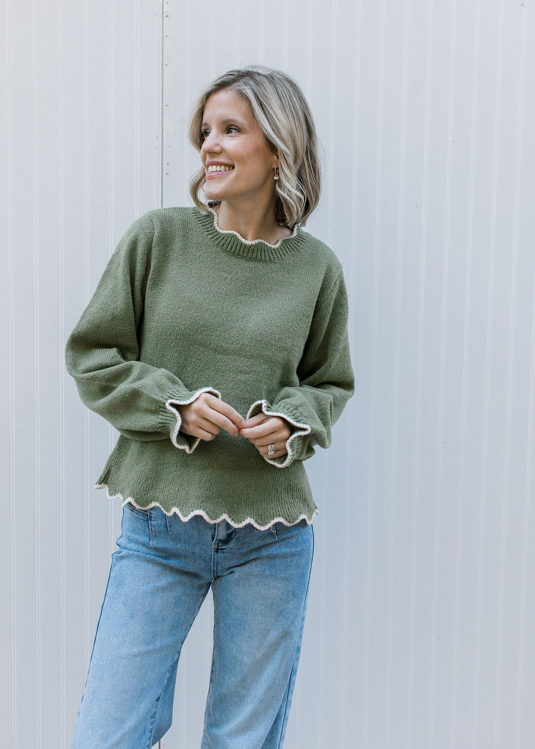 Model wearing jeans and an olive peplum sweater with cream scalloped trim and long sleeves.