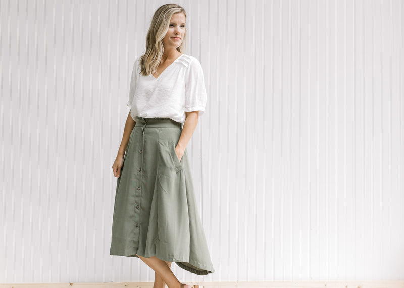 Model wearing a white top with an olive midi skirt, with button up closure and pockets. 
