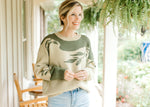 Model wearing a sage sweater with olive tree detail, long sleeves and ribbed neck, hem and cuff.