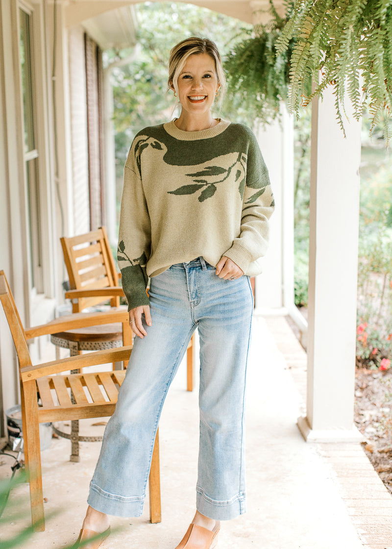 Model wearing jeans with a sage sweater with olive tree detail and long sleeves. 