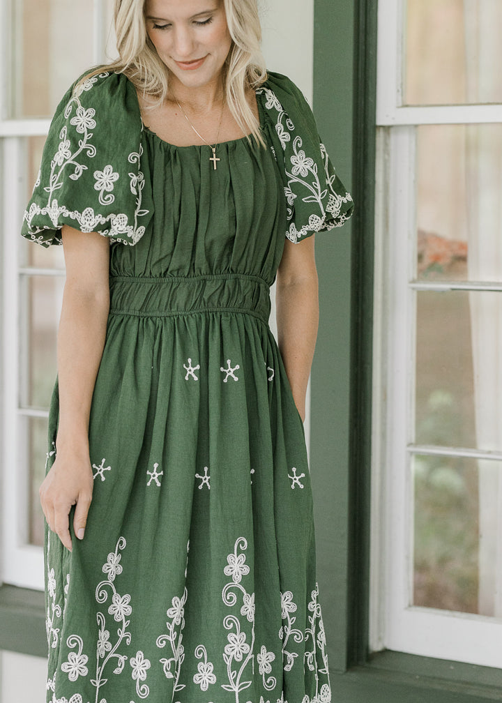 Model wearing a hunter green dress with a square neck, cream floral and pleated bust. 