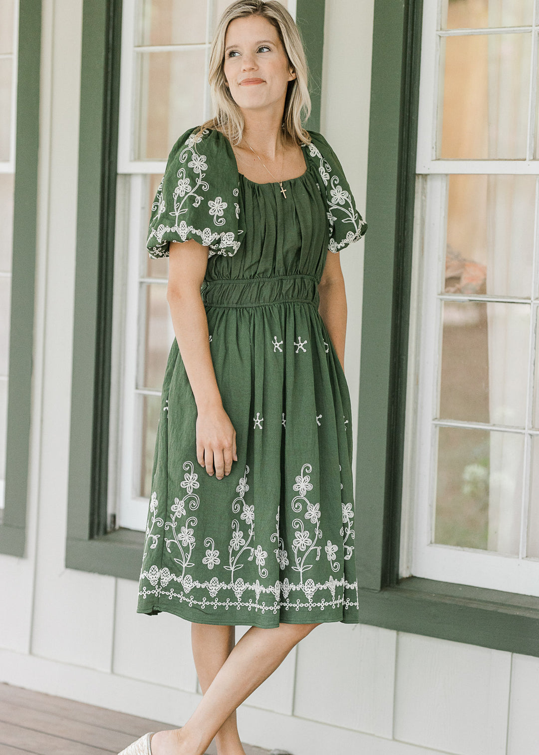 Woman wearing a green dress with white floral embroidery standing in front of a window.