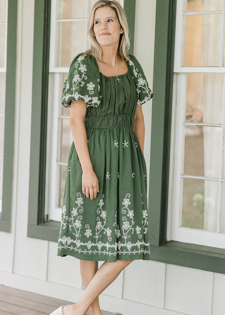 Woman wearing a green dress with white floral embroidery standing in front of a window.