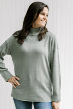 Model wearing an olive long sleeve sweater with a turtleneck, drop shoulder and small side slits. 