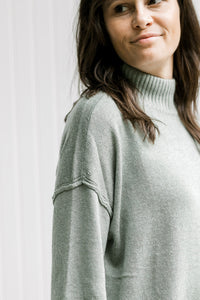 Close up of drop shoulder and turtleneck on a model in an olive colored long sleeve sweater. 