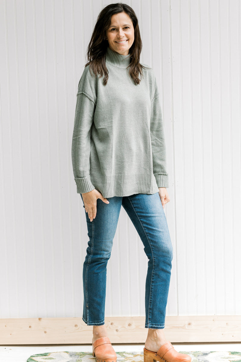 Model wearing jeans with an olive long sleeve sweater with a turtleneck and small side slits. 