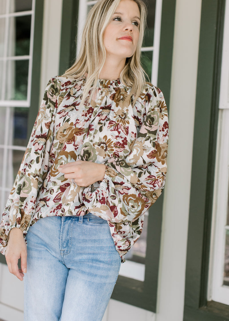 Model wearing jeans and a cream top with warm fall floral, high neck ruffle and long sleeves.