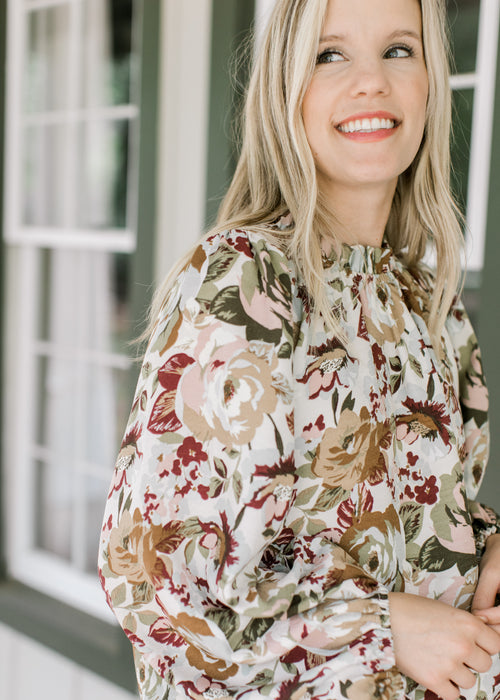 Model wearing a cream top with maroon, pink and green floral, high ruffle neck and long sleeves.