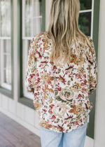 Back view of model wearing a cream top with warm fall floral, high neck ruffle and long sleeves.   