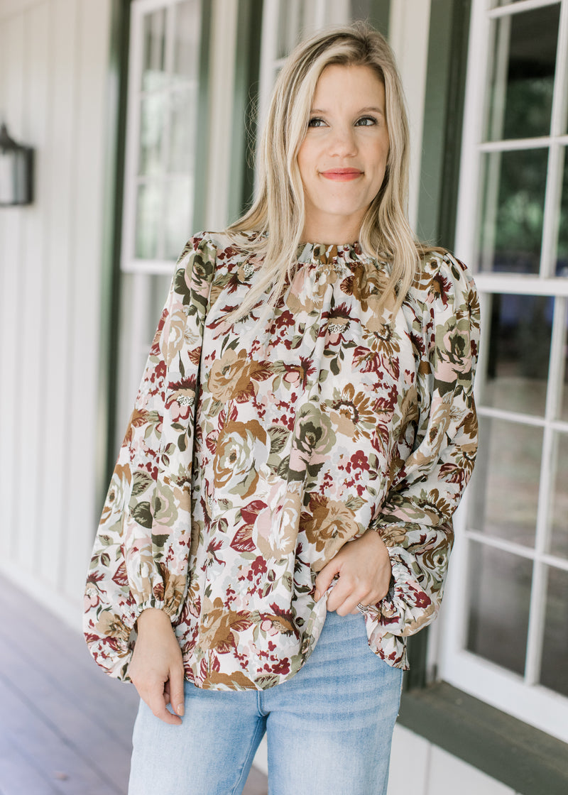 Model wearing a cream top with warm fall floral, high neck ruffle and long sleeve with elastic cuff.