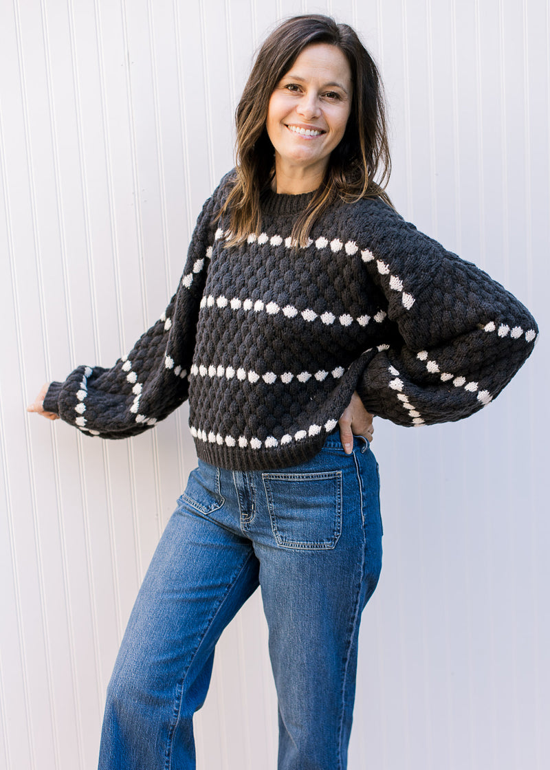 Model wearing jeans and a black textured sweater with white striped dots and long sleeves.