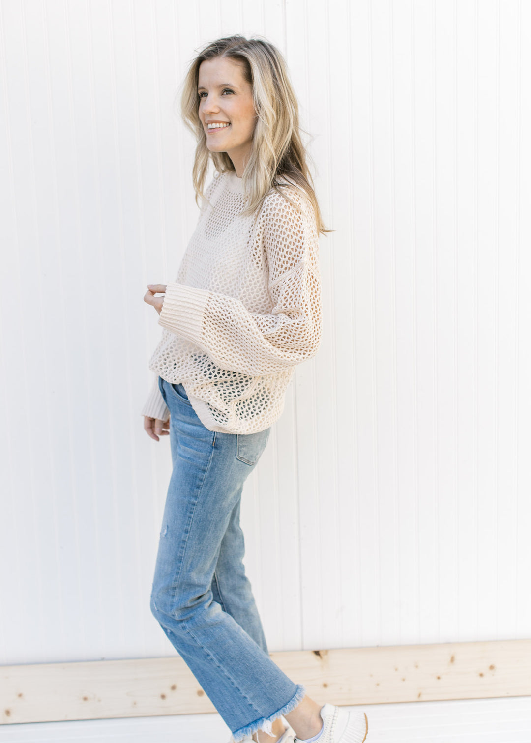 Model wearing jeans, sneakers and a cream open weave sweater with a round neck and long sleeves.