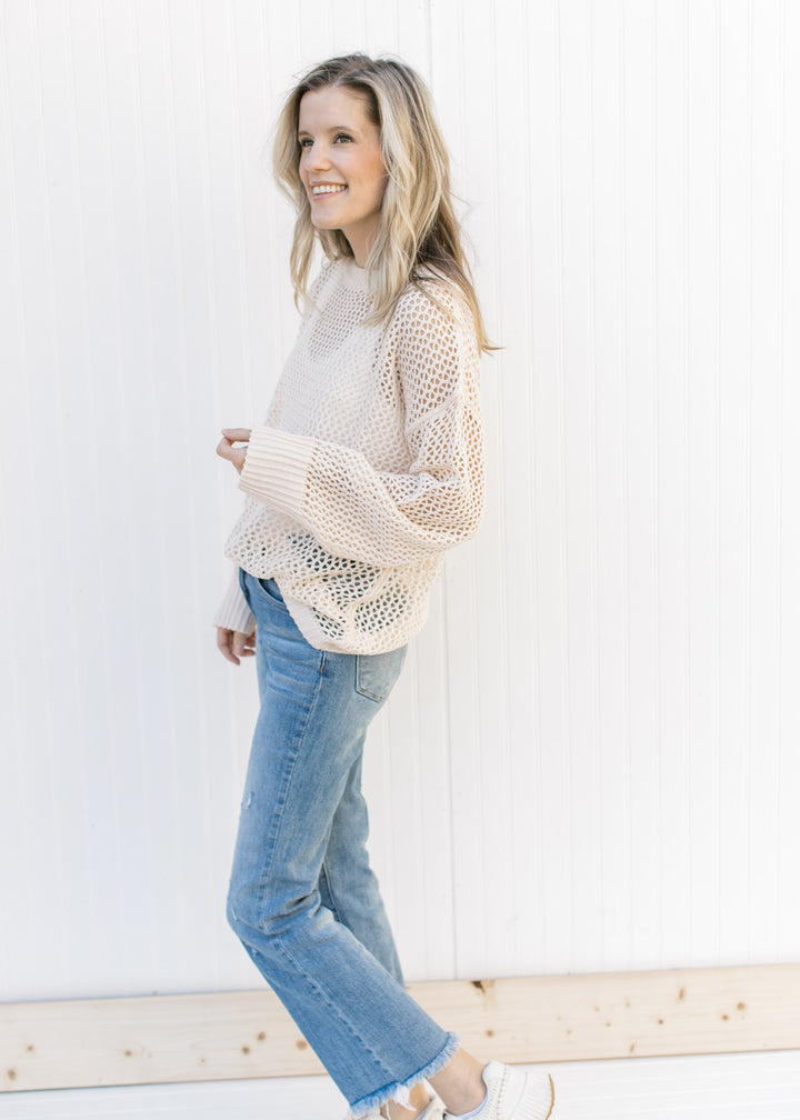 Model wearing jeans, sneakers and a cream open weave sweater with a round neck and long sleeves.