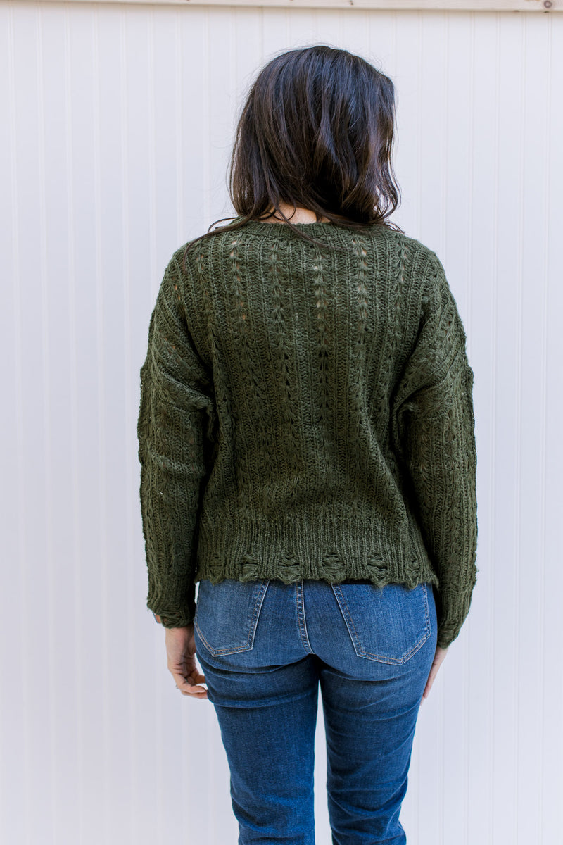 Back view of Model wearing an olive cable knit sweater with a distressed hem and cuff. 