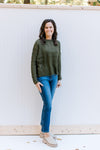 Model wearing jeans and mules with an olive cable knit sweater with a distressed hem and cuff. 