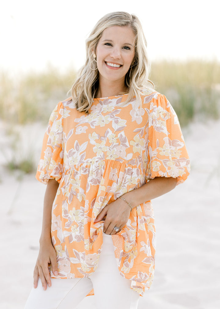 Model wearing a orange top with a neutral floral patter, short puff sleeves and a babydoll fit. 