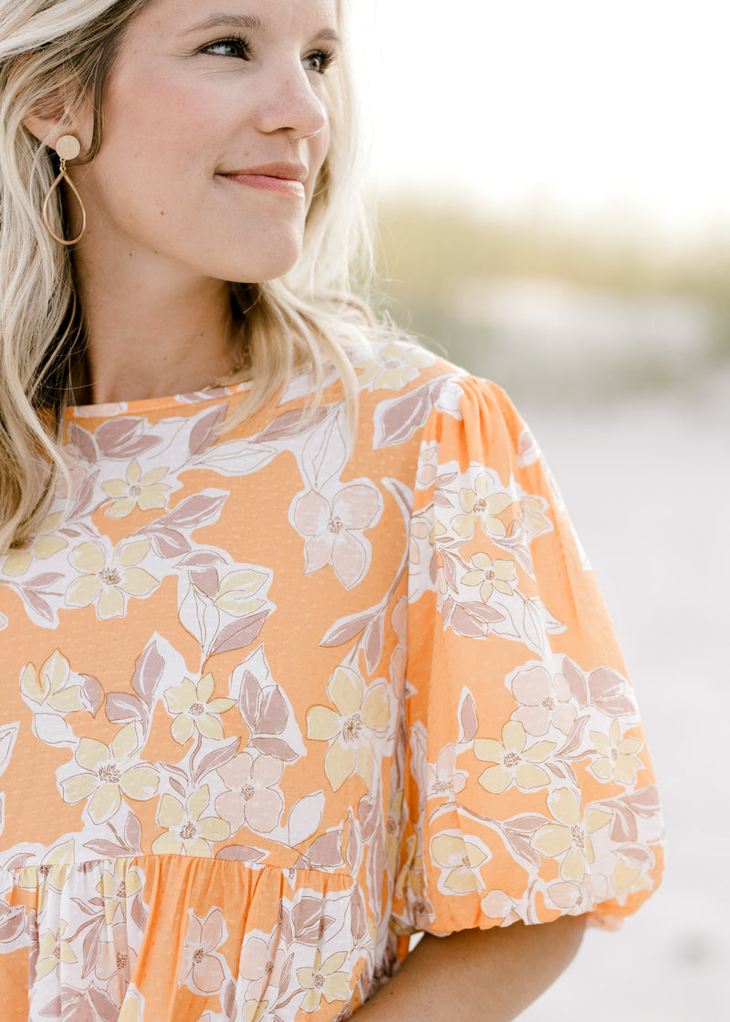 Close up view of short puff sleeves and round neck on an orange top with a neutral floral pattern. 