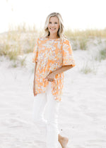 Model wearing white jeans with an orange top with a neutral floral patter and short puff sleeves. 