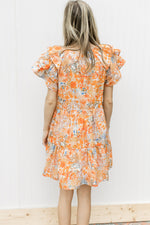 Back view of Model wearing a cream dress with orange and blue flowers and short sleeves.