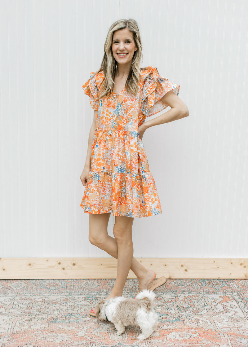 Model wearing sandals with a cream dress with orange and blue flowers and layered short sleeves.